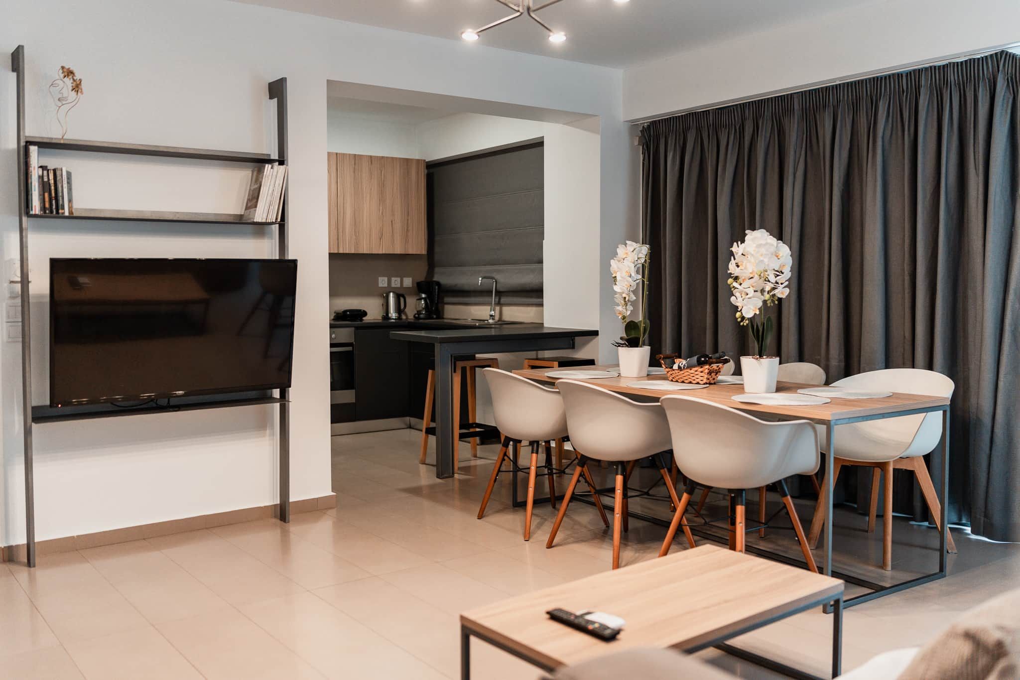 Uma sala de estar moderna com televisão de tela plana, uma mesa de jantar com cadeiras e uma cozinha ao fundo.