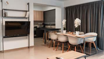 Uma sala de estar moderna com televisão de tela plana, uma mesa de jantar com cadeiras e uma cozinha ao fundo.