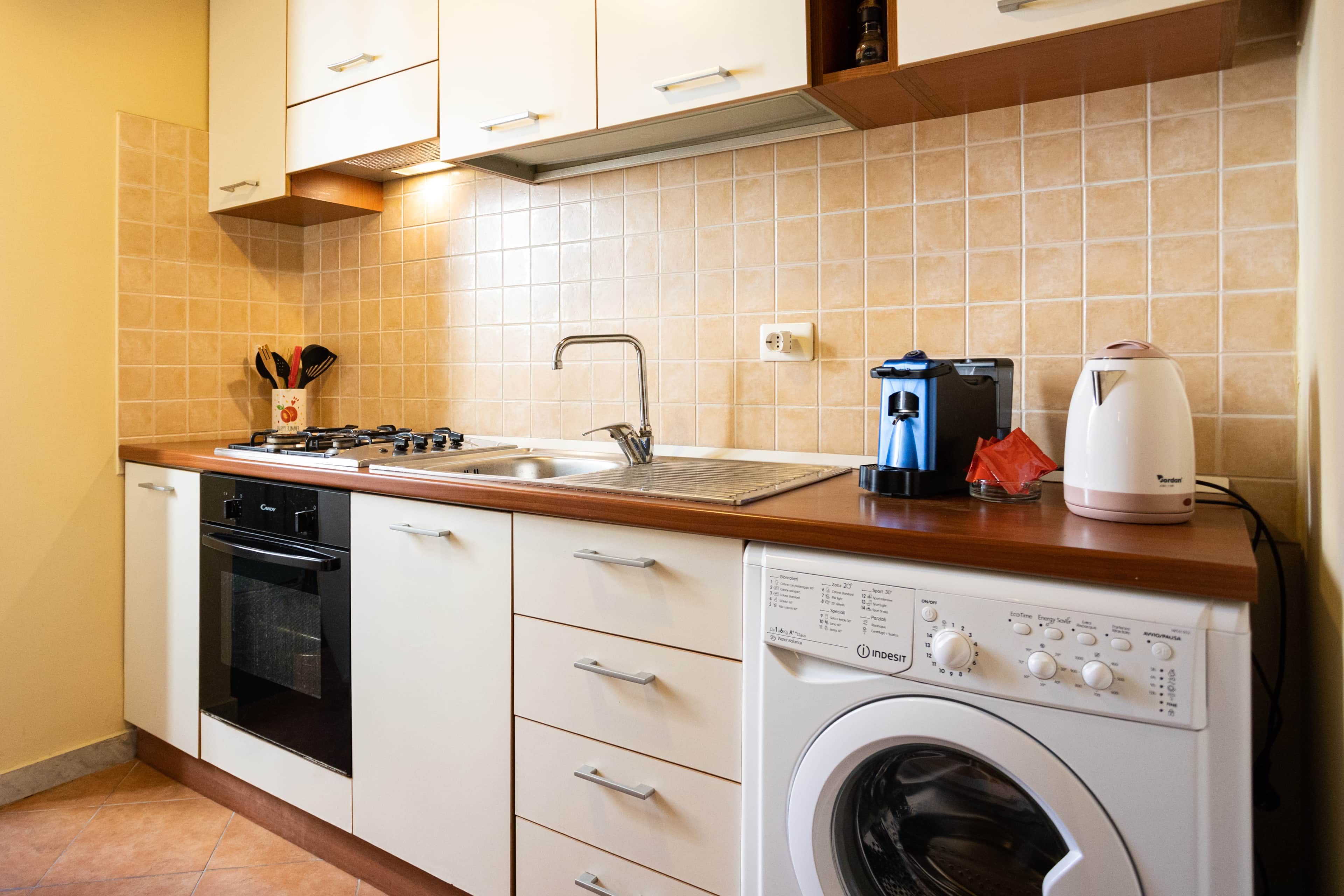 Une cuisine compacte équipée d’un four encastré, d’un lave-linge, d’un évier et d’une machine à café.