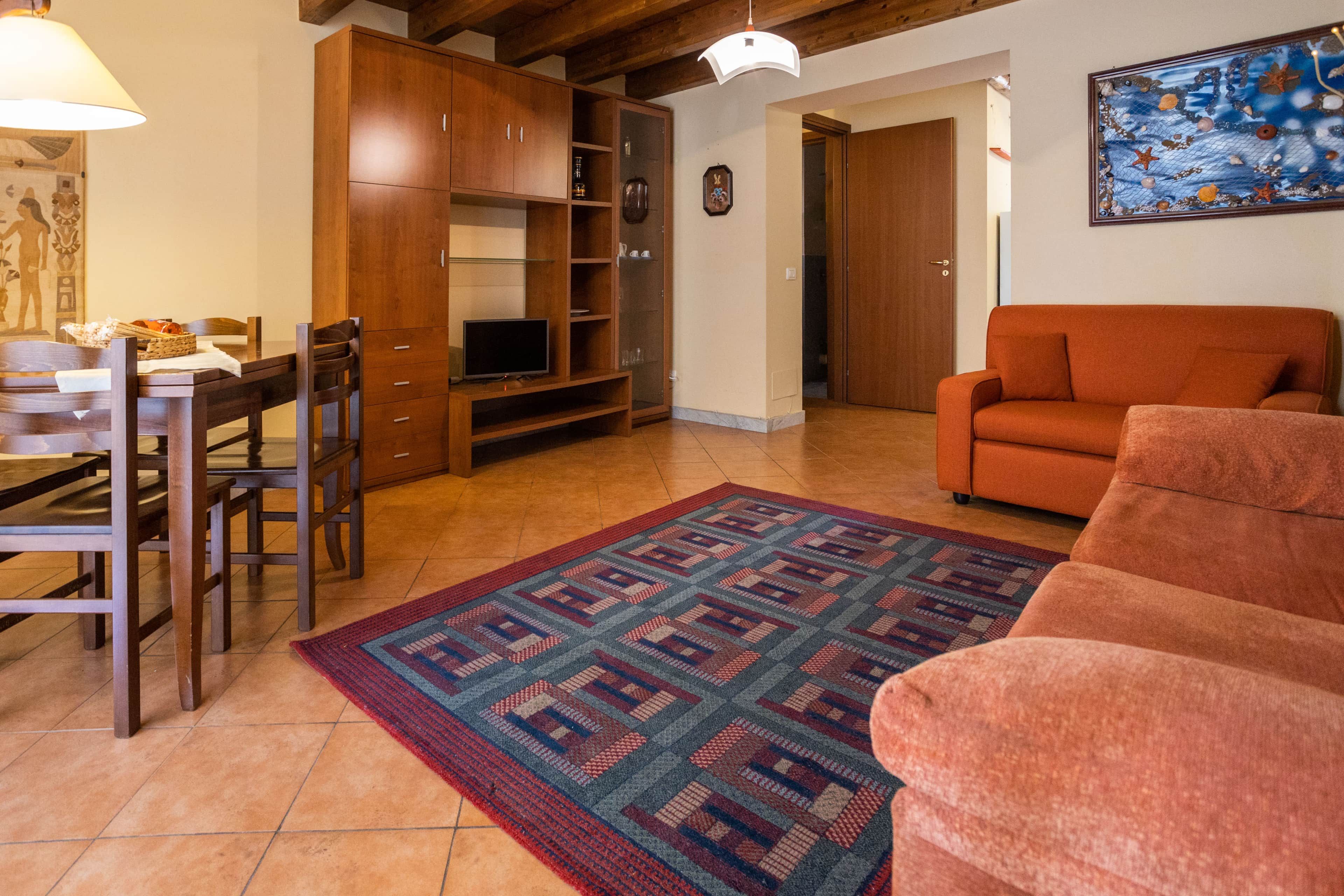 Un salon comprenant un canapé, un téléviseur posé sur un meuble, une table à manger avec des chaises et un tapis à motifs.