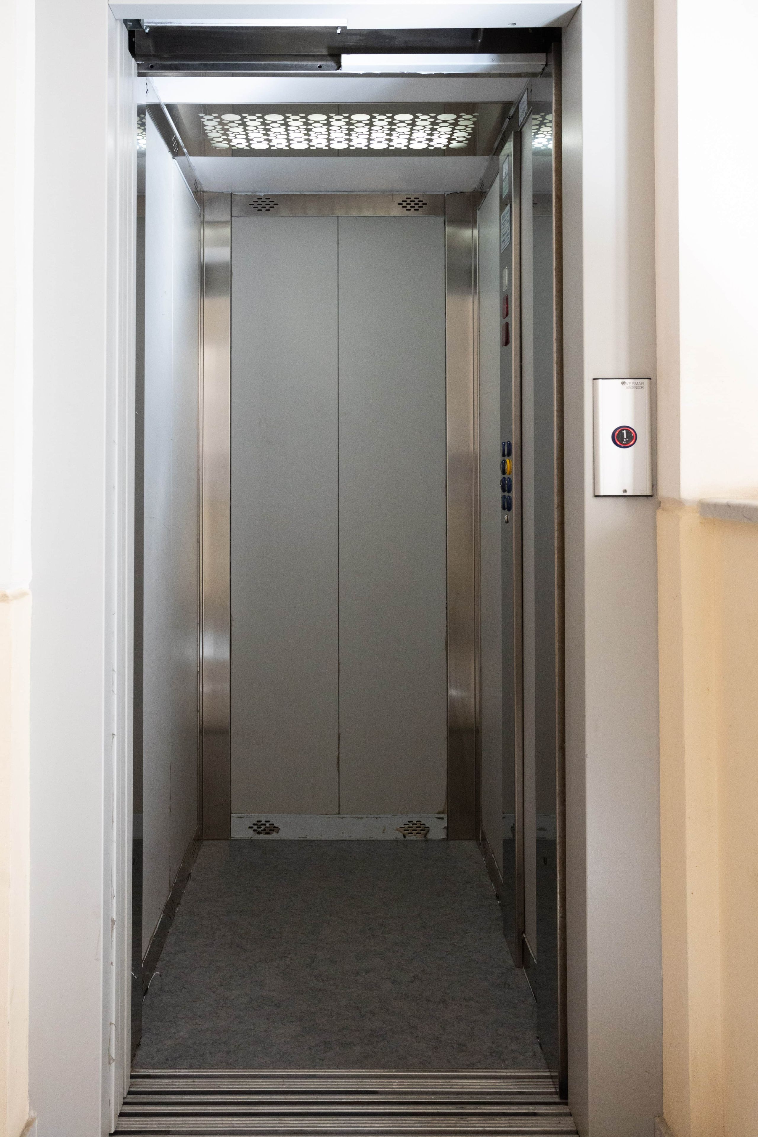 Un ascenseur à l’intérieur métallique, avec un panneau de commande sur le côté droit et un sol recouvert de moquette.