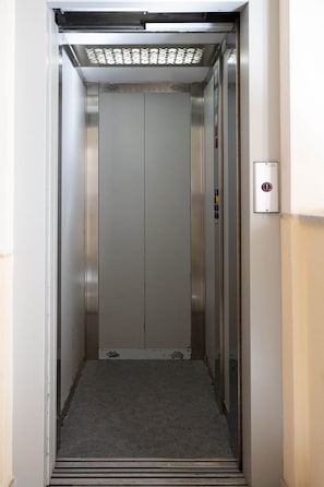 Un ascenseur à l’intérieur métallique, avec un panneau de commande sur le côté droit et un sol recouvert de moquette.