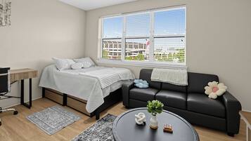 A modern living room with a bed, a black sofa, a coffee table with a plant, and a window with a view of buildings.