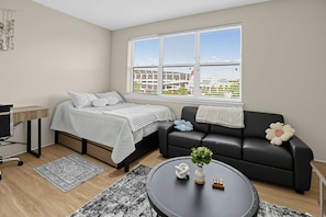 Una sala de estar moderna con una cama, un sofá negro, una mesa de centro con una planta y un ventanal con vistas a edificios.