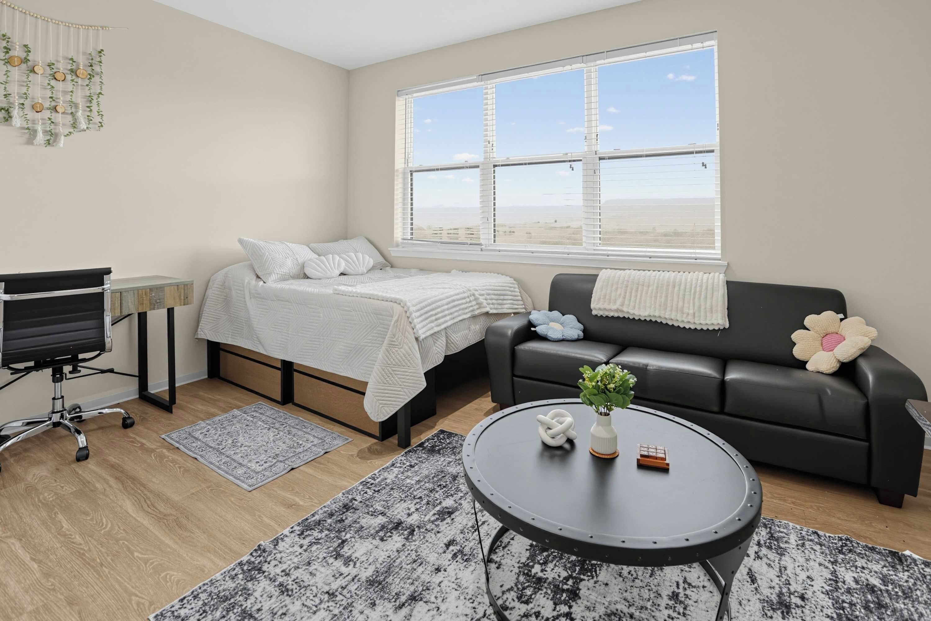A modern bedroom with a bed, a black leather sofa, a round coffee table, and a desk with a chair.