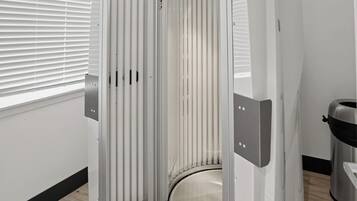 A room with a corner shower enclosure, a window with blinds, and a trash bin.