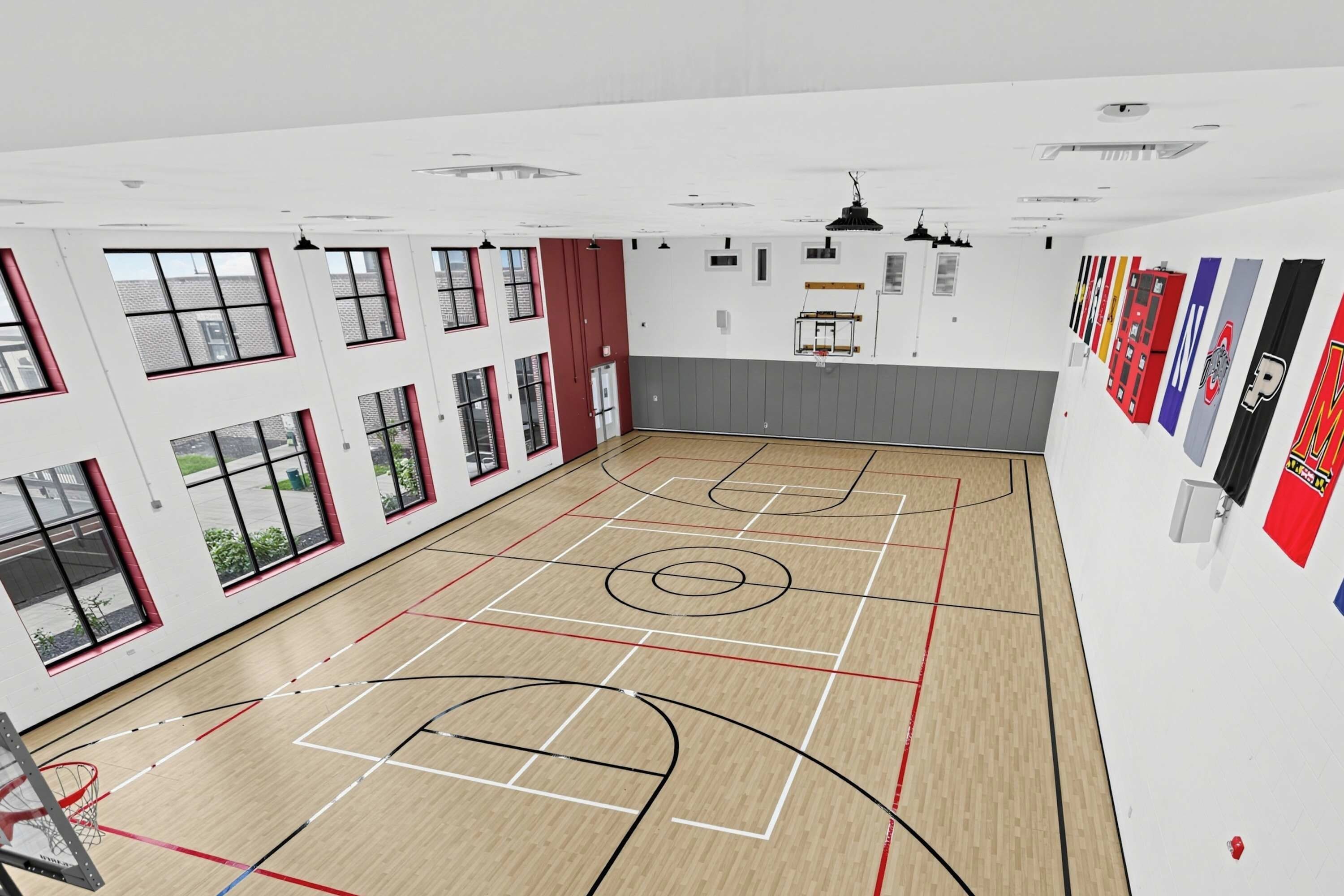 A basketball court with a wooden floor, multiple windows with red frames, and a basketball hoop.