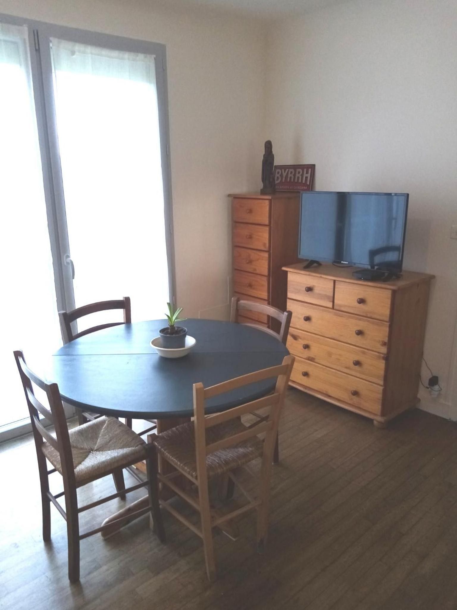 Un comedor con una mesa redonda, dos sillas, una pequeña planta en maceta y un mueble de madera con cajones.