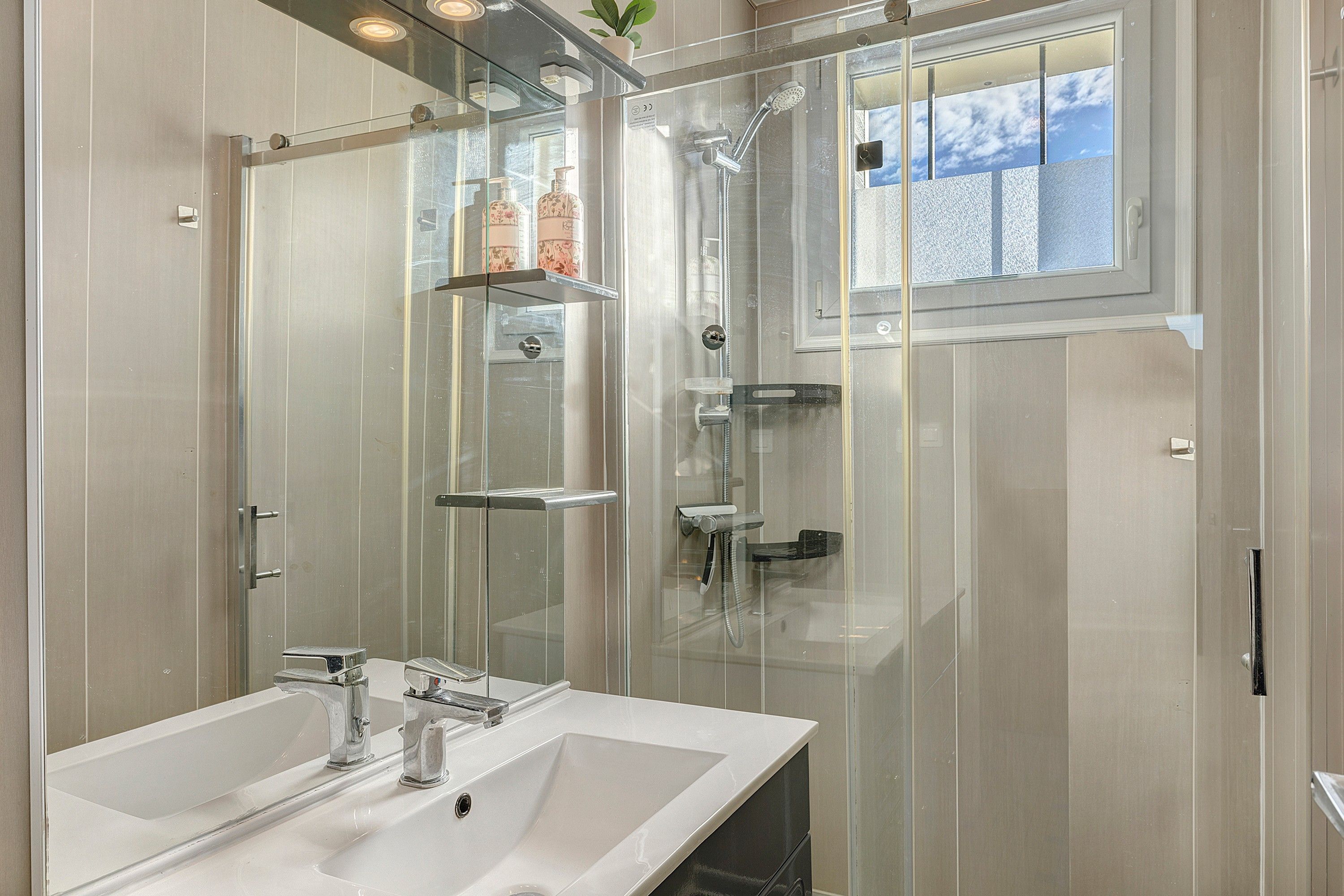 A modern bathroom with a glass-enclosed shower, a white sink, and a window providing natural light.