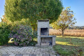 A stone-built outdoor fireplace with a firebox, surrounded by lush greenery and a clear sky.