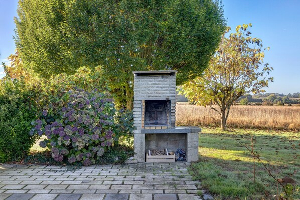 A stone-built outdoor fireplace with a firebox, surrounded by lush greenery and a clear sky.
