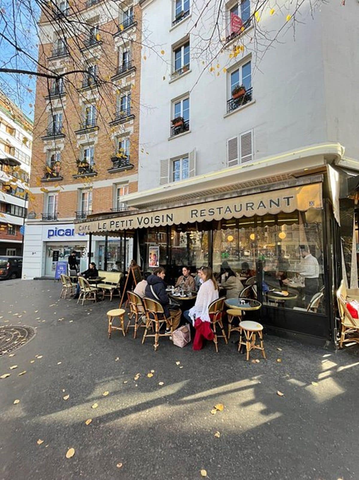 Un café al aire libre con sillas y mesas de mimbre, una tienda con un letrero que dice 'LE PETIT VOISIN RESTAURANT', y un edificio con varias ventanas y balcones al fondo.