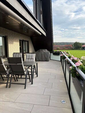 Een buitenterras met een tafel, stoelen, een overdekte barbecue en uitzicht op een groen veld.