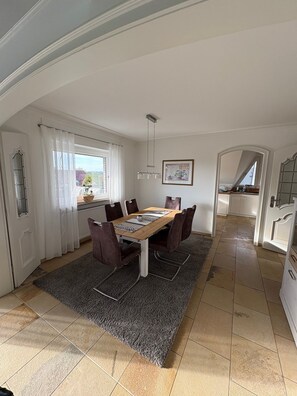 Een eetruimte met een tafel, stoelen en een vloerkleed. Er is een raam met gordijnen, een schilderij aan de muur en een keuken in de achtergrond zichtbaar.