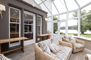 A cozy indoor seating area with wicker sofas and a wooden bench, large windows offering a view of a lake and trees. - Luxury country house with outdoor hot tub overlooking Loch Ard & Ben Lomond (Aberfoyle)