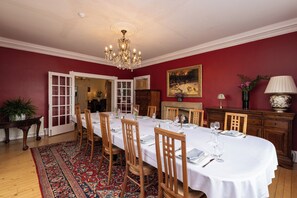 A dining room with a long table set for six, wooden chairs, a chandelier, a red wall, and a painting. - Luxury country house with outdoor hot tub overlooking Loch Ard & Ben Lomond (Aberfoyle)