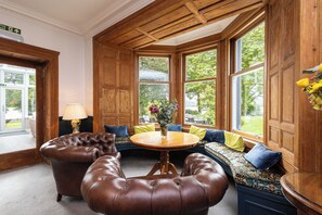 Quarto com mesa redonda de madeira, poltronas de couro e um banco com almofadas. Janelas amplas oferecem vista para o verde lá fora.