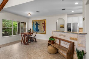 A dining area with a table and chairs, a large painting on the wall, a kitchen with a refrigerator, and a stone fireplace. - Private Hill Country Retreat 3-bedroom house with Resort Pool (Austin)