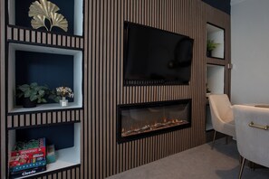 Een moderne woonkamer met een flatscreen televisie die aan een houten wand bevestigd is, een open haard eronder en planken met gezelschapsspellen en decoratieve objecten.