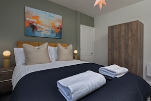 A neatly made bed with pillows and a blanket, a wooden wardrobe, a painting on the wall, and a nightstand with a lamp. - The Clifton Townhouse, 5Bed with Hot Tub, King Bed (Scarborough)