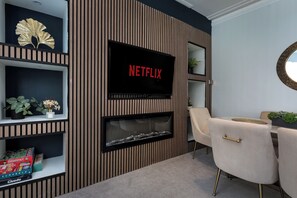 Een moderne woonkamer met een flatscreen-tv waarop 'NETFLIX' staat, een open haard en een zithoek met een ronde tafel en stoelen.