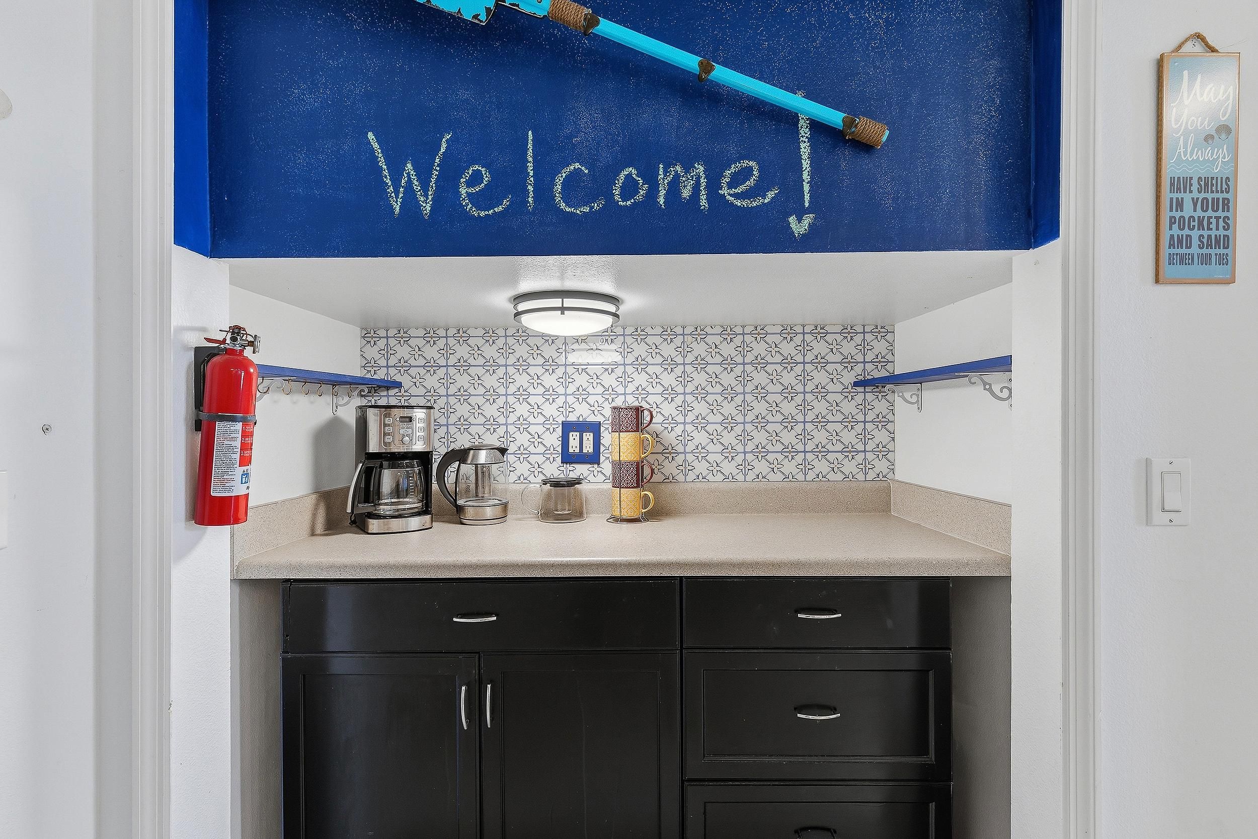 Una cocina con un cartel de 'Welcome', una cafetera, un hervidor, una taza y un extintor.