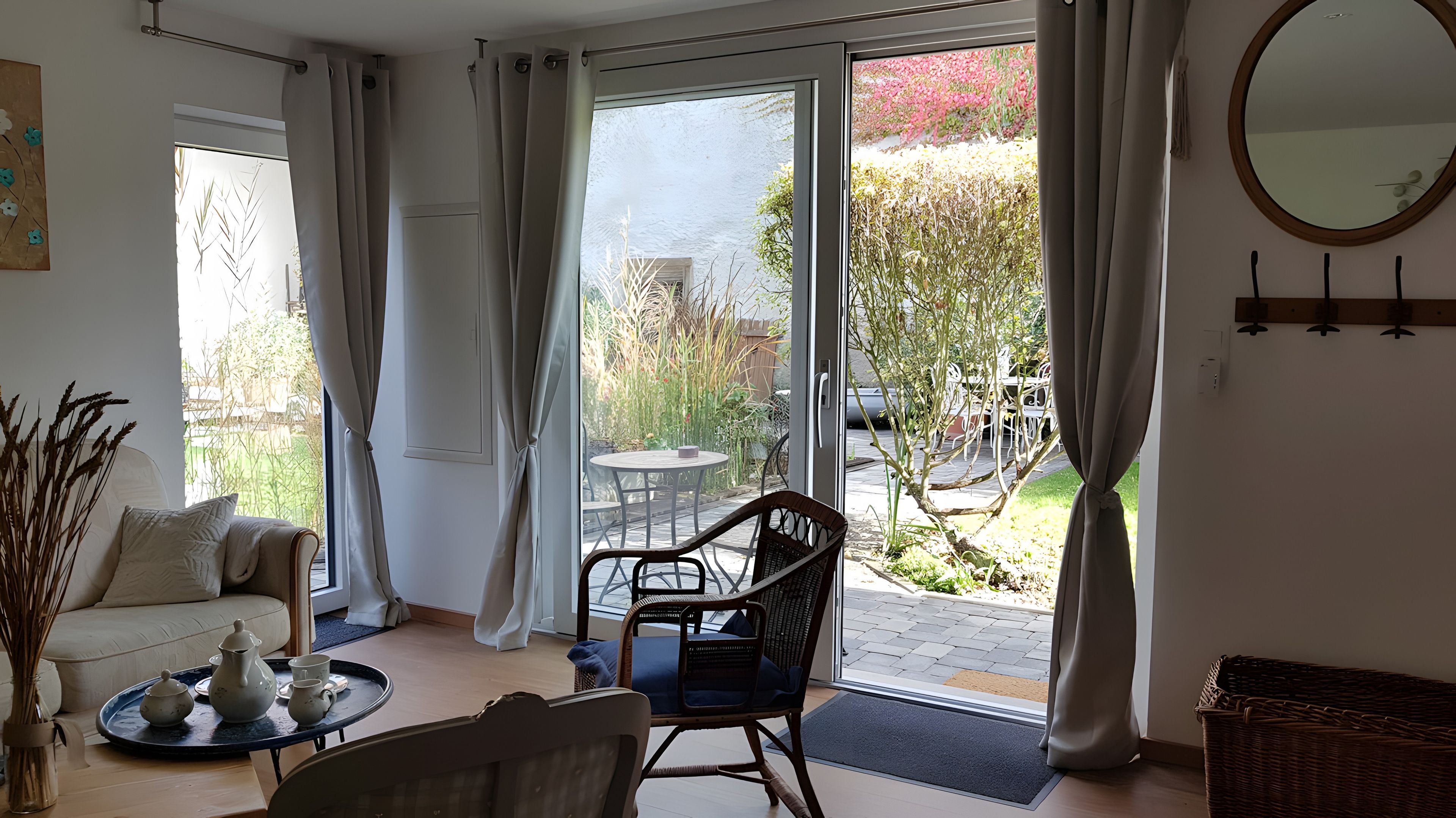 Una habitación con sofá, una mesa de comedor con sillas, una silla de mimbre, una bandeja con teteras y vista a un jardín a través de una puerta de cristal.