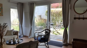 Una habitación con sofá, una mesa de comedor con sillas, una silla de mimbre, una bandeja con teteras y vista a un jardín a través de una puerta de cristal.