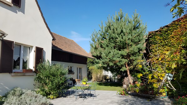 Ein Wohnhaus mit einer gepflasterten Terrasse, einem Esstisch mit Stühlen, einem großen immergrünen Baum und verschiedenen Kübelpflanzen.