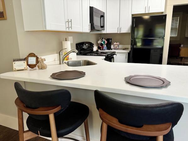 A kitchen with white cabinets, a stainless steel sink, a microwave, a stove, and a refrigerator. There are two black chairs with wooden frames.