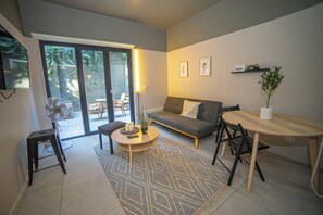 A modern living room with a sofa, a coffee table, and a dining table. There is a sliding glass door leading to an outdoor area with a chair and a table. - 1-Bedroom Apartment + Terrace · Featured Design (H04) (Providencia)