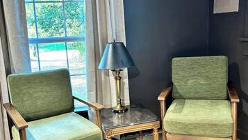 Dos sillones tapizados en verde con patas de madera, una mesita con una lámpara y una ventana con cortinas.