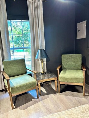 Dos sillones tapizados en verde con patas de madera, una mesita con una lámpara y una ventana con cortinas.