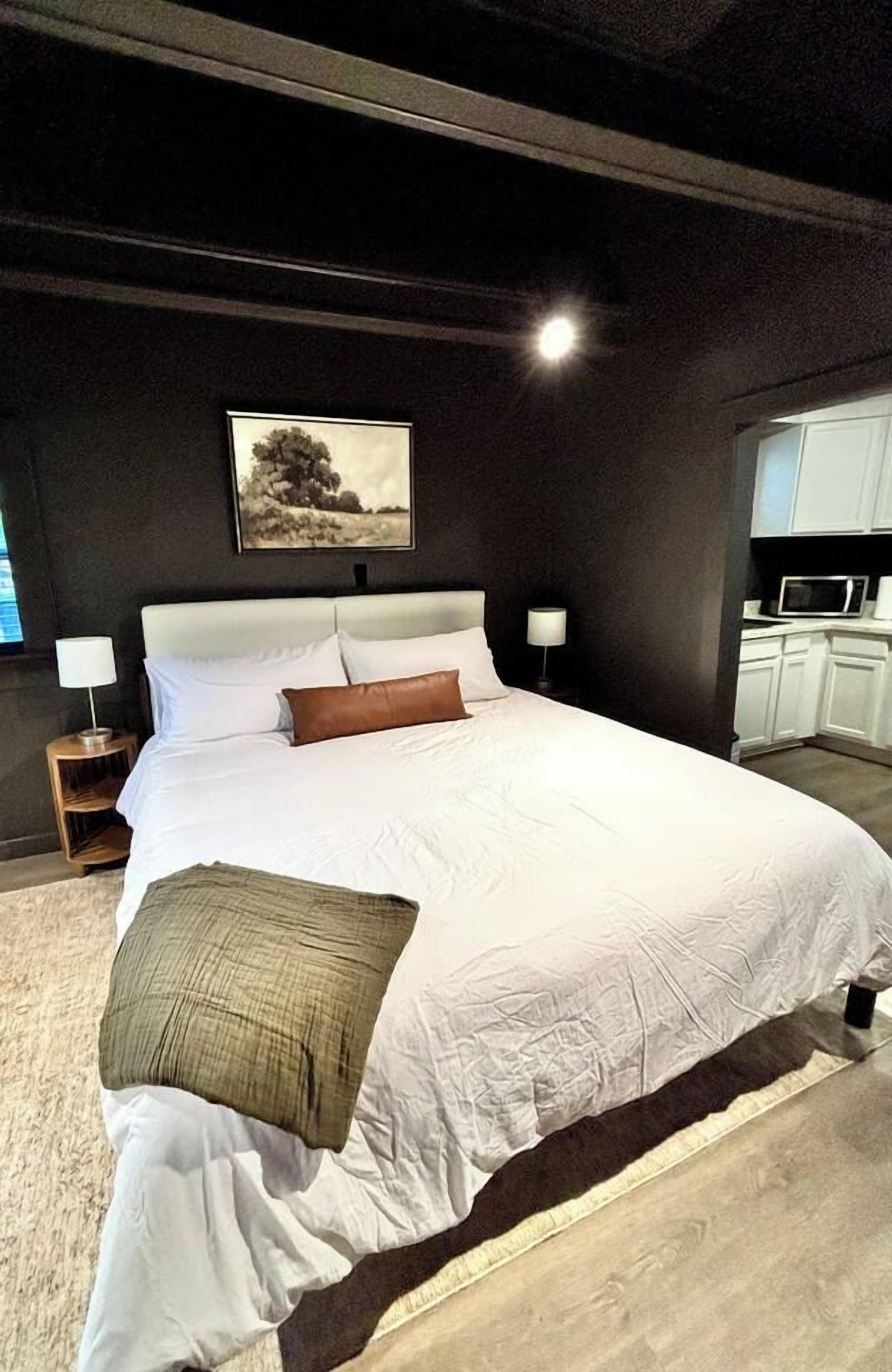 A bedroom with a large bed, bedside lamps, a painting on the wall, and a kitchenette in the background.