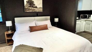 A bedroom with a large bed, bedside lamps, a painting on the wall, and a kitchenette in the background.