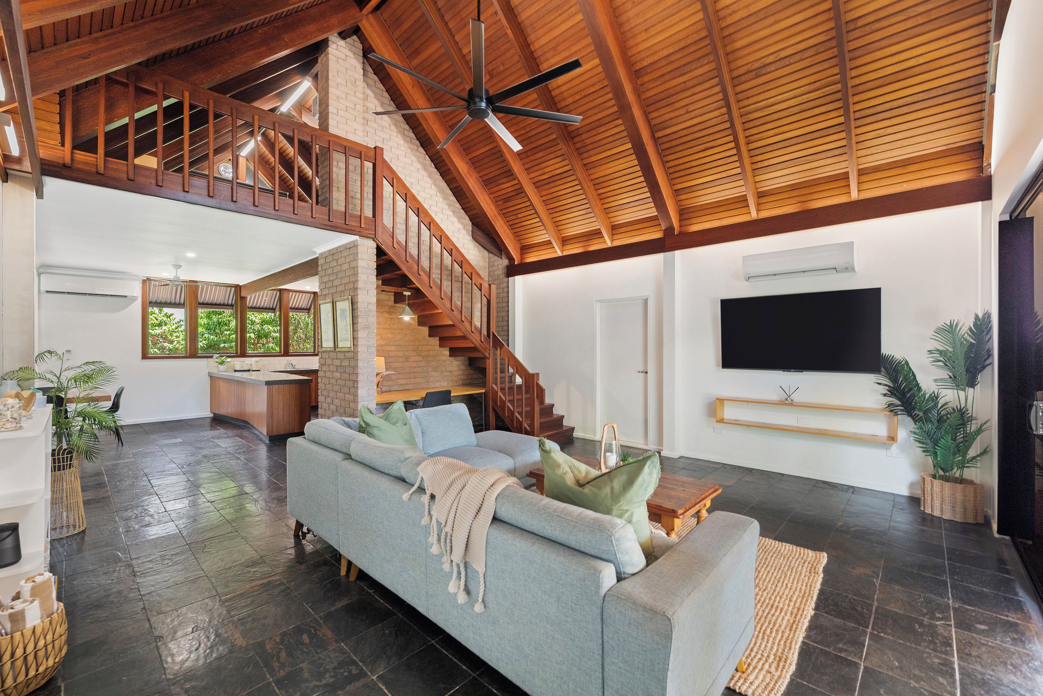 A spacious living area with a high ceiling, a staircase, a large sofa, and a flat-screen TV mounted on the wall.