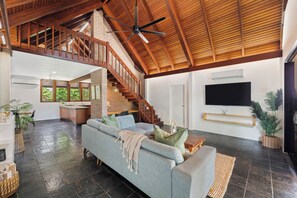 A spacious living area with a high ceiling, a staircase, a large sofa, and a flat-screen TV mounted on the wall.