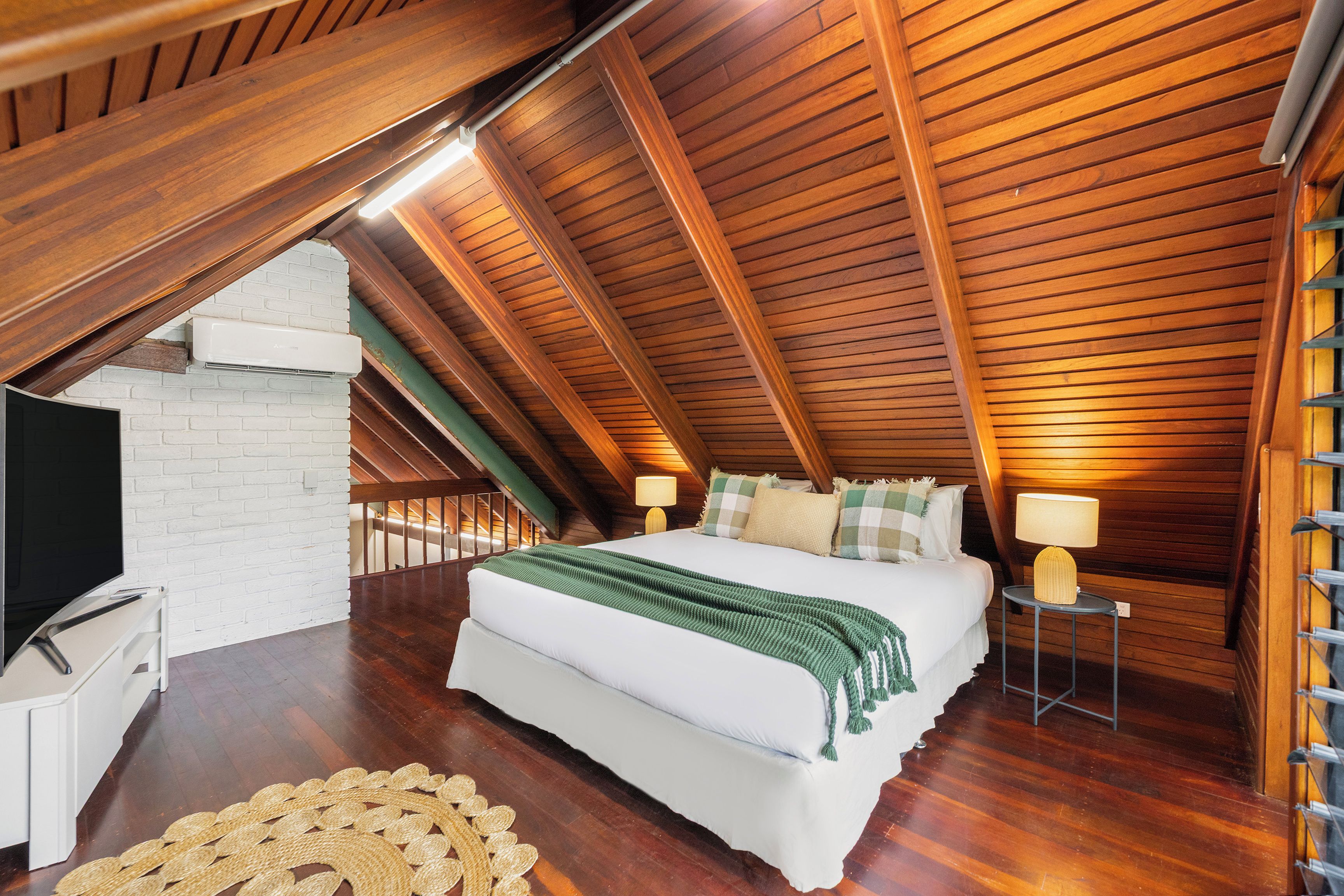 Ein Schlafzimmer mit schrägverdeckter Decke, einem Bett, Nachttischlampen, einem Fernseher und einer gewobenen Fußmatte.