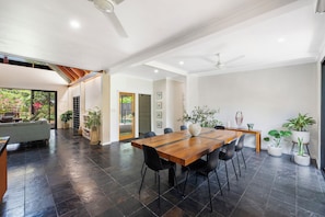 A spacious living area with a large wooden dining table, black chairs, and a grey sofa. There are several potted plants and a ceiling fan. - Serena Oasis Tropical Sanctuary with Private Pool (Nightcliff)