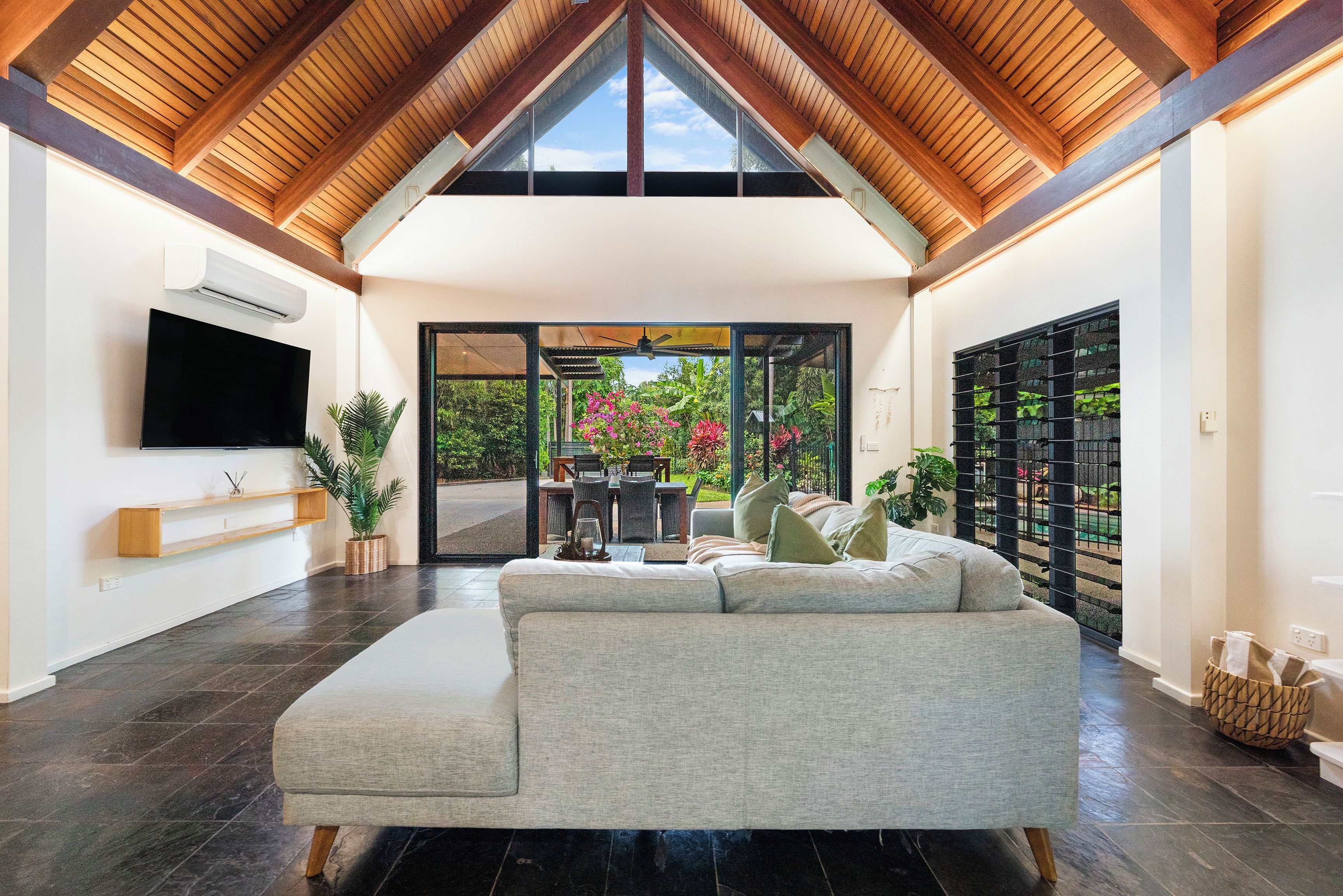 A modern living room with a high ceiling, a flat-screen TV mounted on the wall, a sofa, and a dining area with chairs and a table.