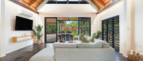 A modern living room with a high ceiling, a flat-screen TV mounted on the wall, a sofa, and a dining area with chairs and a table.