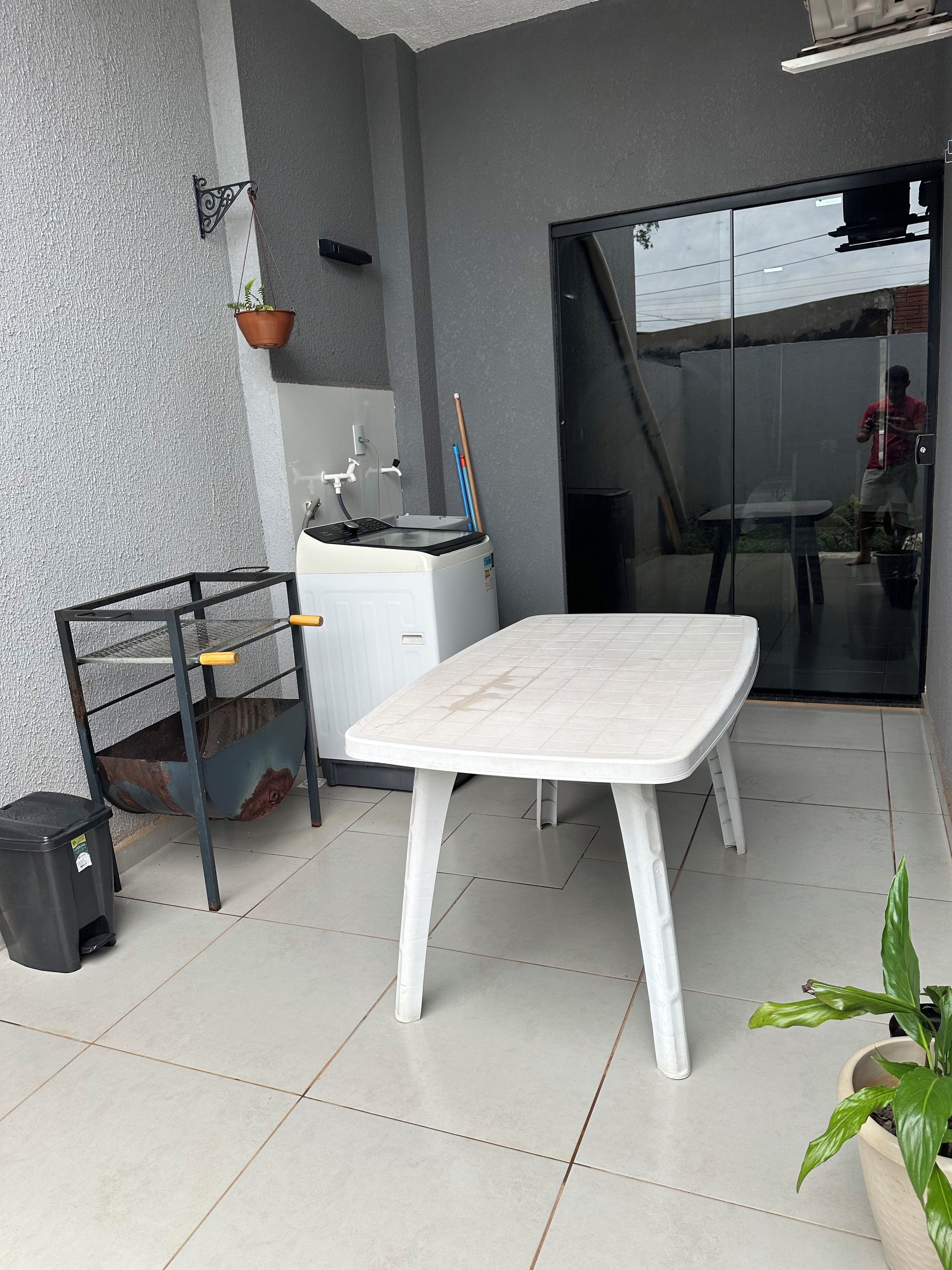 Un pequeño espacio al aire libre con una mesa blanca, un estante metálico, un dispensador de agua y una planta en maceta.