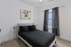 Ein Schlafzimmer mit einem Bett, einem Nachttisch, einer Lampe, einem Fenster mit Vorhängen und einem Gemälde an der Wand.