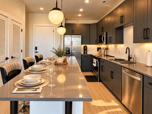 A modern kitchen with a central island, stainless steel appliances, and a dining area with a long table set for a meal. - The Downtown Social | 2BR City Escape (Birmingham)