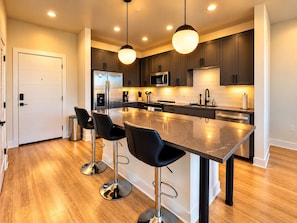 A modern kitchen with a central island, bar stools, stainless steel appliances, and pendant lighting. - The Downtown Social | 2BR City Escape (Birmingham)