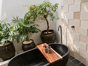 Una bañera negra con una bandeja de madera, rodeada de plantas en macetas y una pared de piedra.