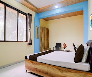 A bedroom with a wooden bed frame, a window with curtains, a chair, a small table with a vase of flowers, and a wardrobe. - Deluxe Hotel Shivneri (Mumbai)