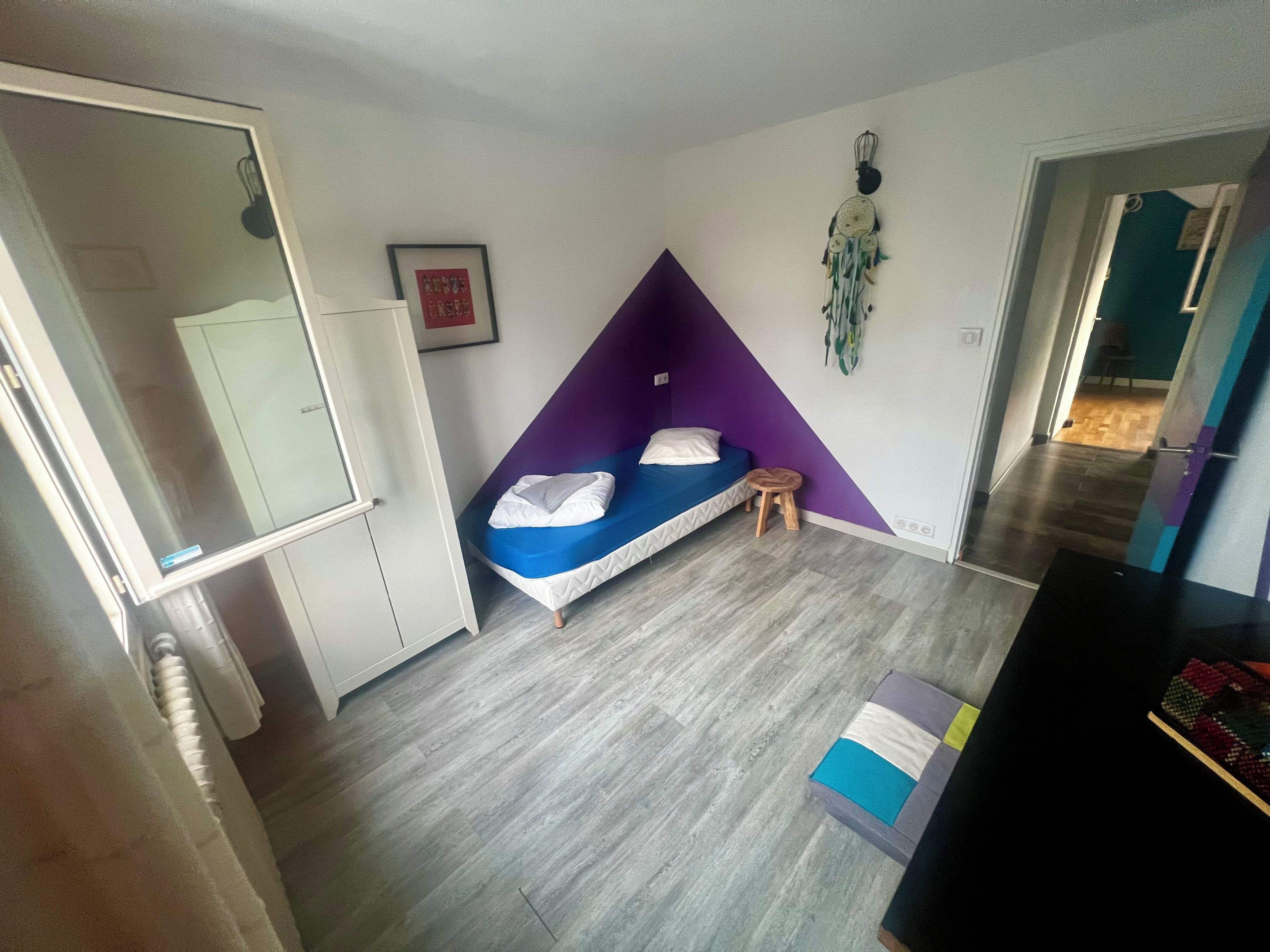 Een kamer met een bed, een klein krukje, een kledingkast en een dreamcatcher aan de muur.