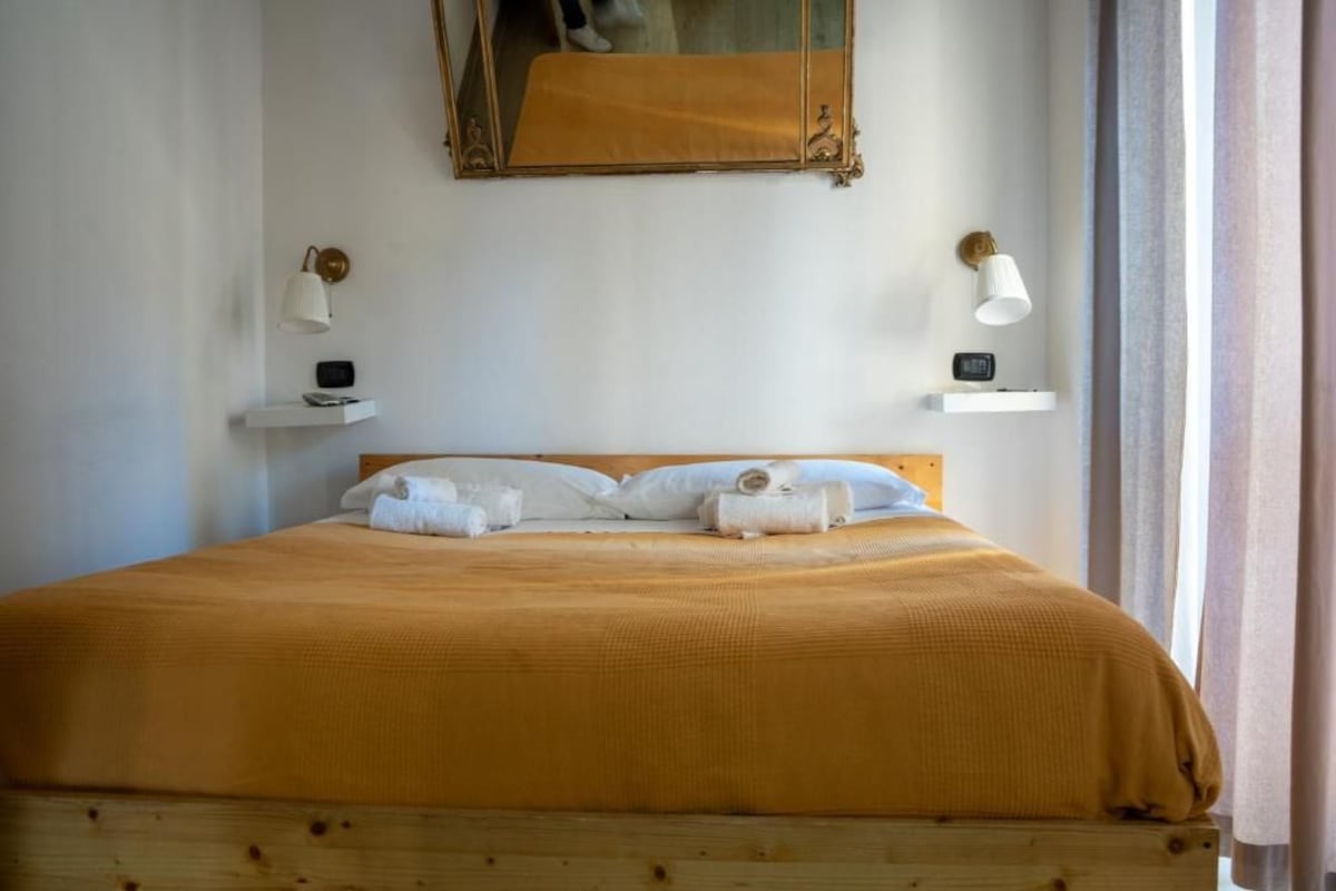 Een bed met een houten hoofdbord, een spiegel erboven en twee nachtkastjes met lampen en kleine elektronische apparaten.