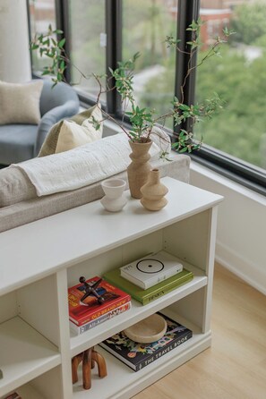 A white bookshelf with books and a small plant, a white table with a vase and a candle, and a window with a view of greenery. - Luxury Designer Collection 519 - Fulton Market (Chicago)